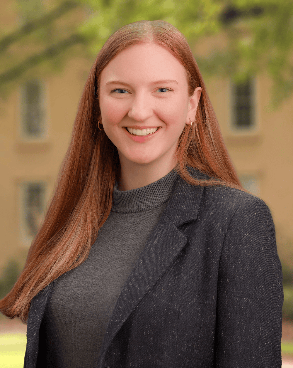 A young woman with long red hair, wearing a dark blazer and gray turtleneck, smiles at the camera. She stands outdoors with a blurred background of trees and a tan building with windows.